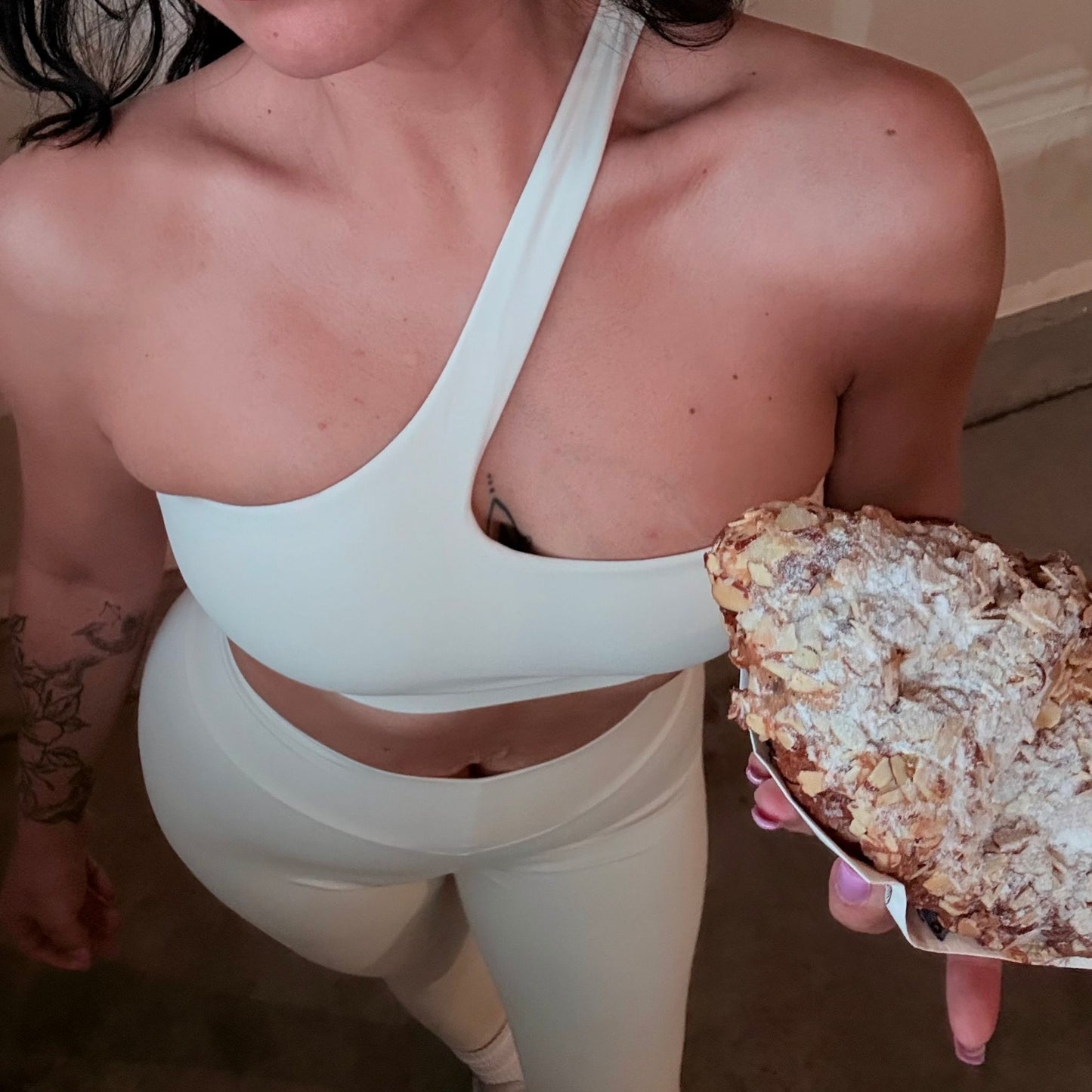 Minimalist cream one-shoulder activewear set from Avvi, featuring a seamless bra and leggings, styled casually while holding a powdered almond croissant.