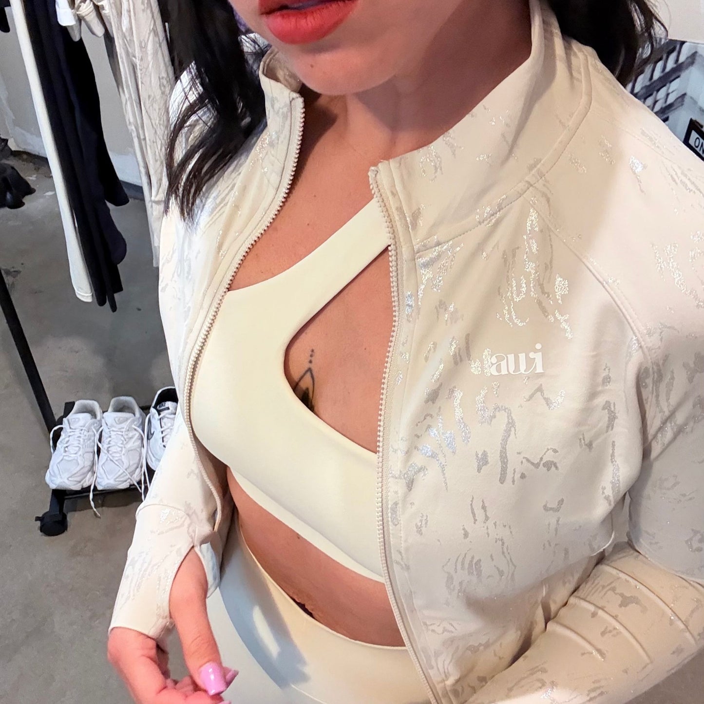 Close-up of cream one-shoulder sports bra layered under a textured cream zip-up jacket with subtle shimmer, featuring the Avvi logo on the chest.