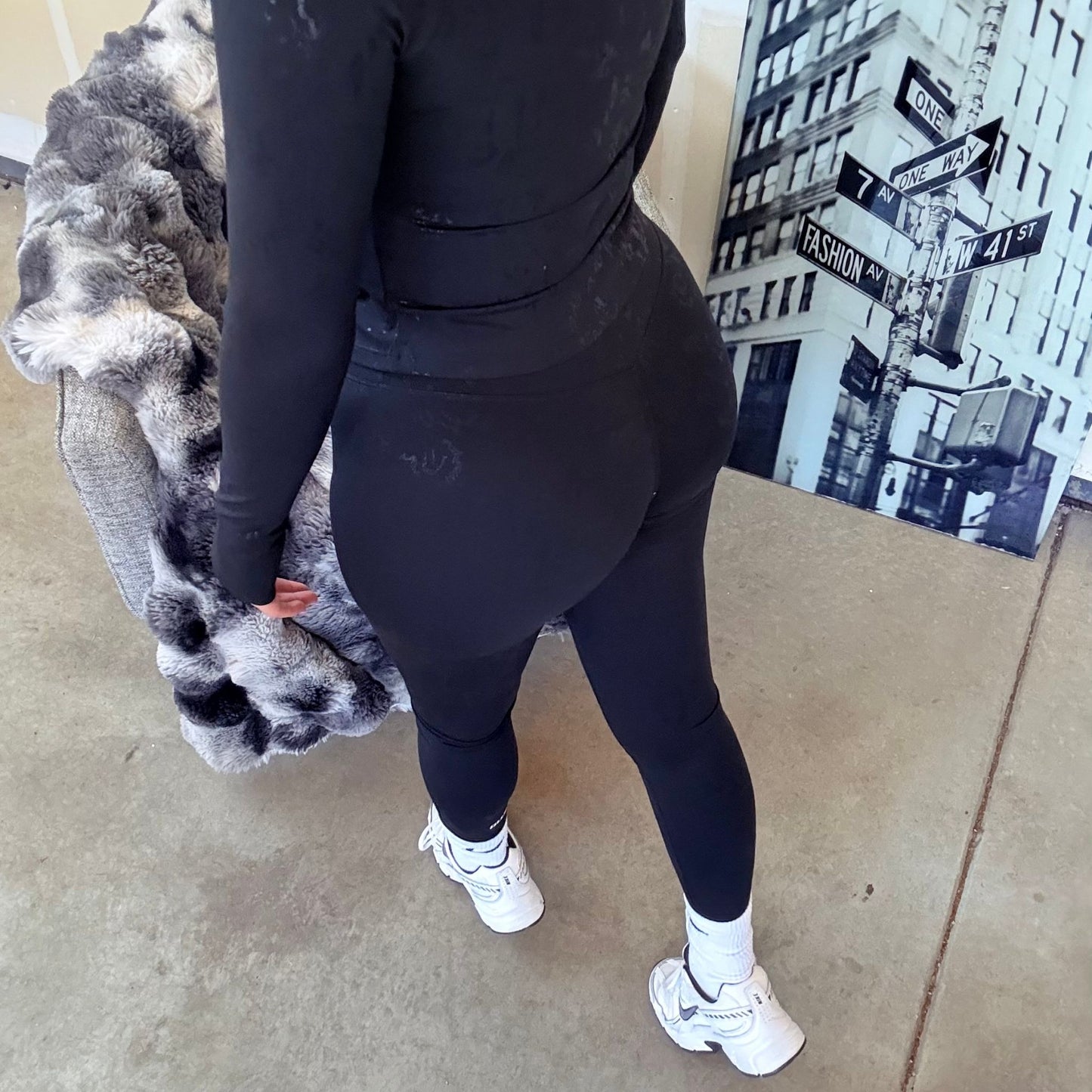 Rear view of a woman in a black textured athletic set, highlighting the curve-enhancing leggings and jacket fit, paired with white sneakers.