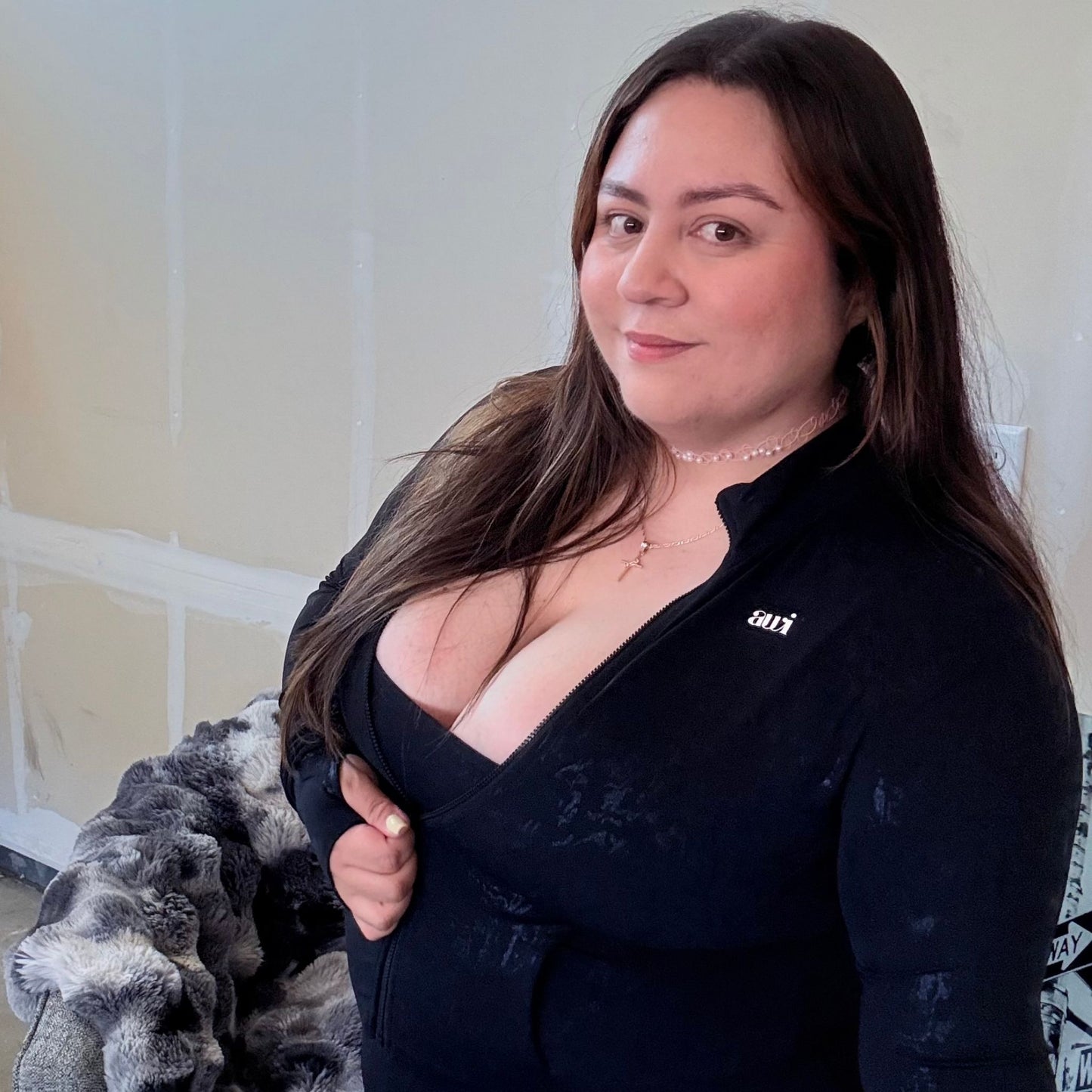 Woman in a black zip-up athletic jacket with subtle glossy texture, smiling confidently while posing indoors.