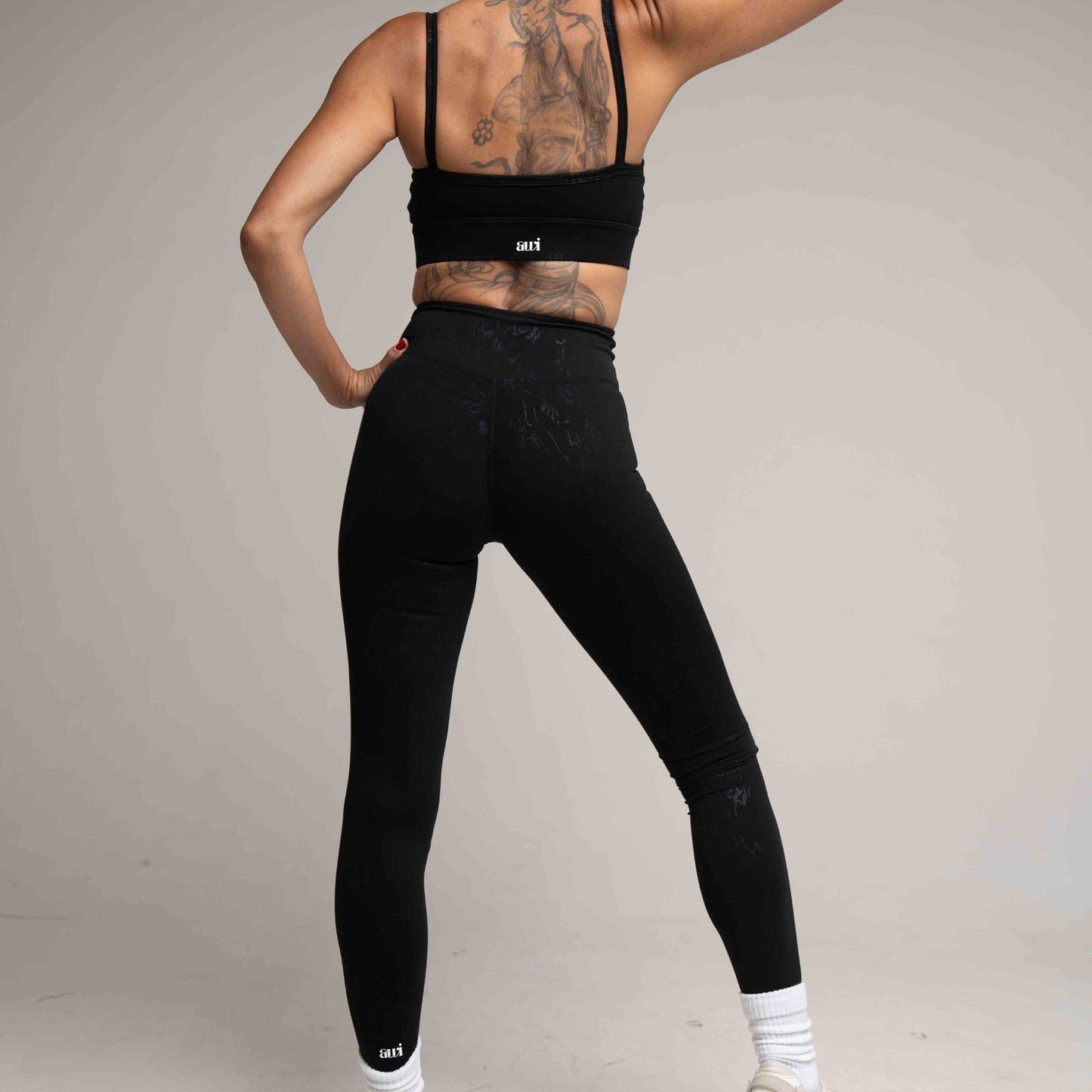 Person wearing black leggings and sports bra with visible tattoos on a plain background