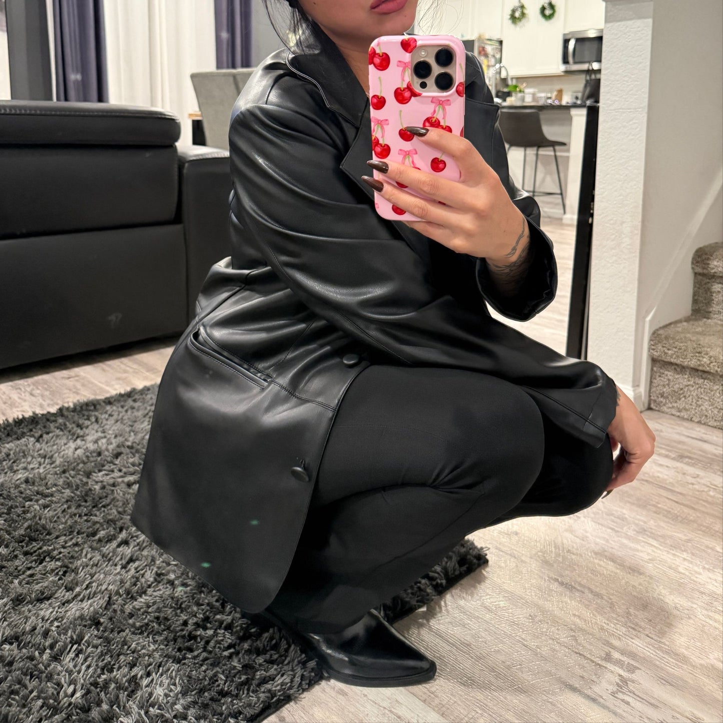 Person wearing an all black outfit, leather coat with black unitard and black boots, taking a mirror selfie in a living room.