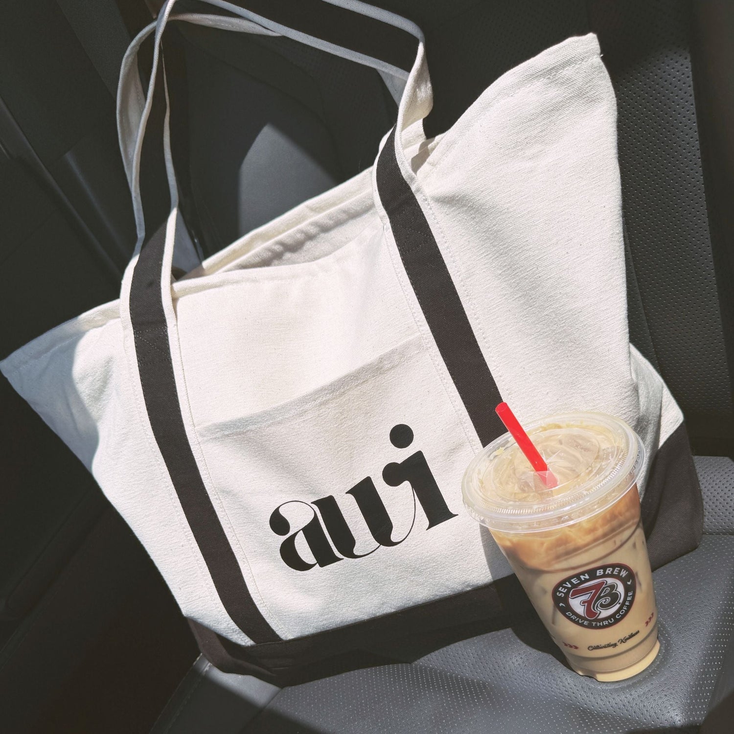 Avvi canvas tote bag with bold black logo and iced coffee on a car seat — minimal, on-the-go lifestyle