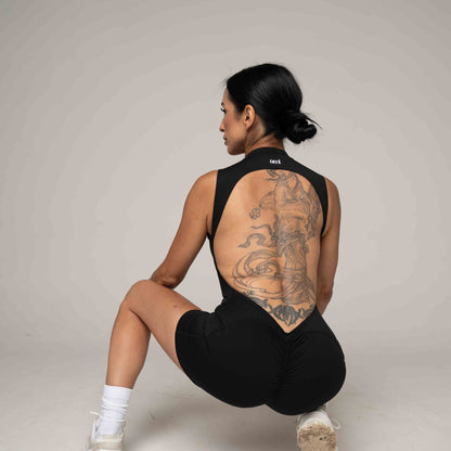 Person wearing a black sleeveless top and shorts with visible tattoos on a plain background