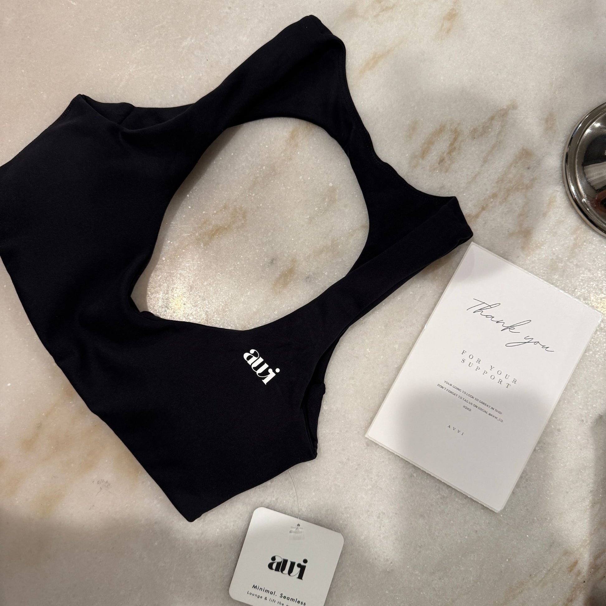Black tank top with 'avvi' branding on a marble surface with a card and packaging.