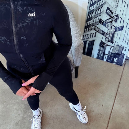 Front view of fitted black zip-up jacket with subtle texture detail and thumbholes, paired with black leggings and white sneakers from Avvi's Espresso Mood collection.